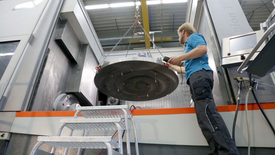 Auf dem Bild ist zu sehen, wie ein Arbeiter eine große, runde Metallplatte mit einem Kran in eine Maschine hebt.